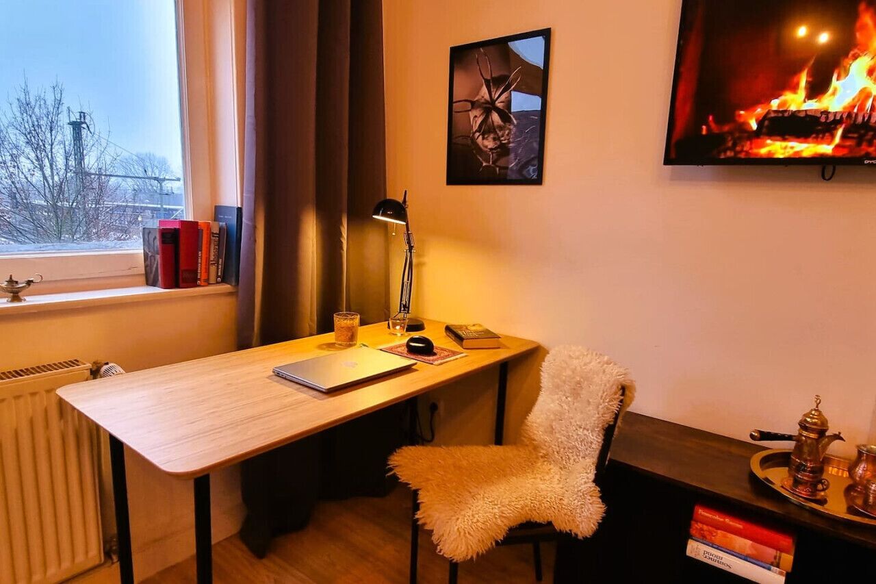 A cozy room with a wooden desk, a chair, a radiator, a window with books on the sill, a lamp, a painting, and a fireplace.