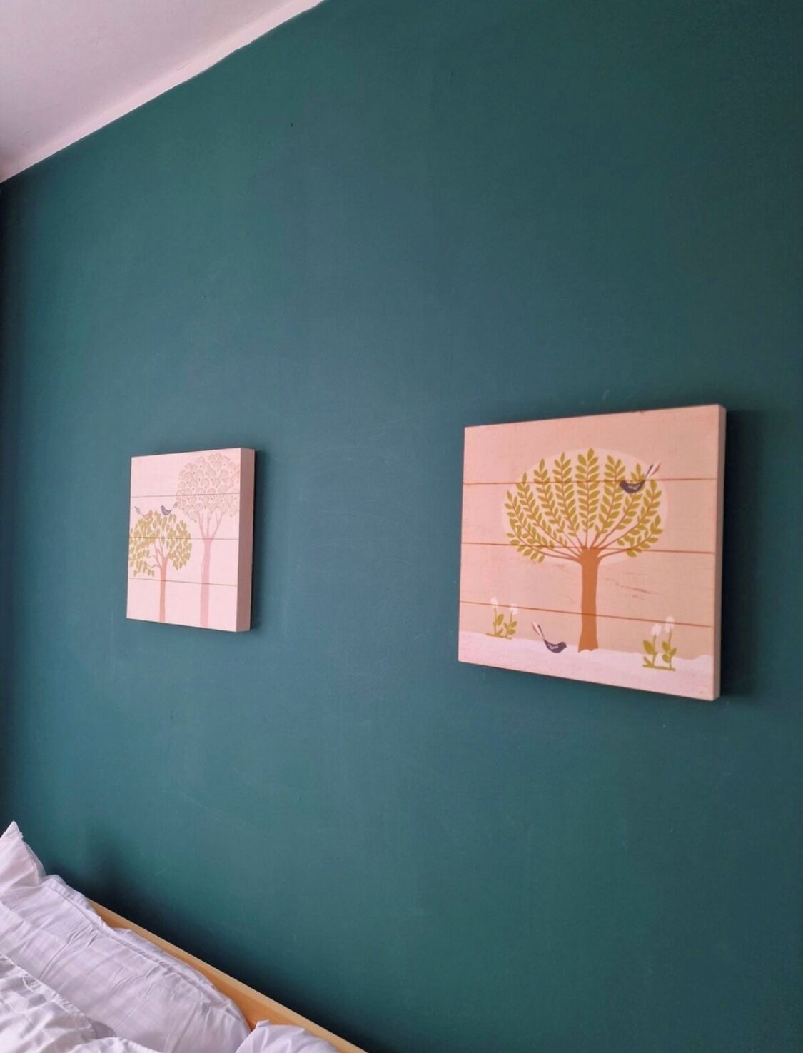 A bedroom with a teal wall, two framed artworks depicting trees, a wooden bed, and a white pillow.