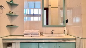 A bathroom with a white sink, teal cabinets, and a mirror above it. There are shelves with toiletries and a window above the mirror. - Vacation home with beautiful sea view near Guidaloca, Scopello and the Zingaro (Castellammare del Golfo)