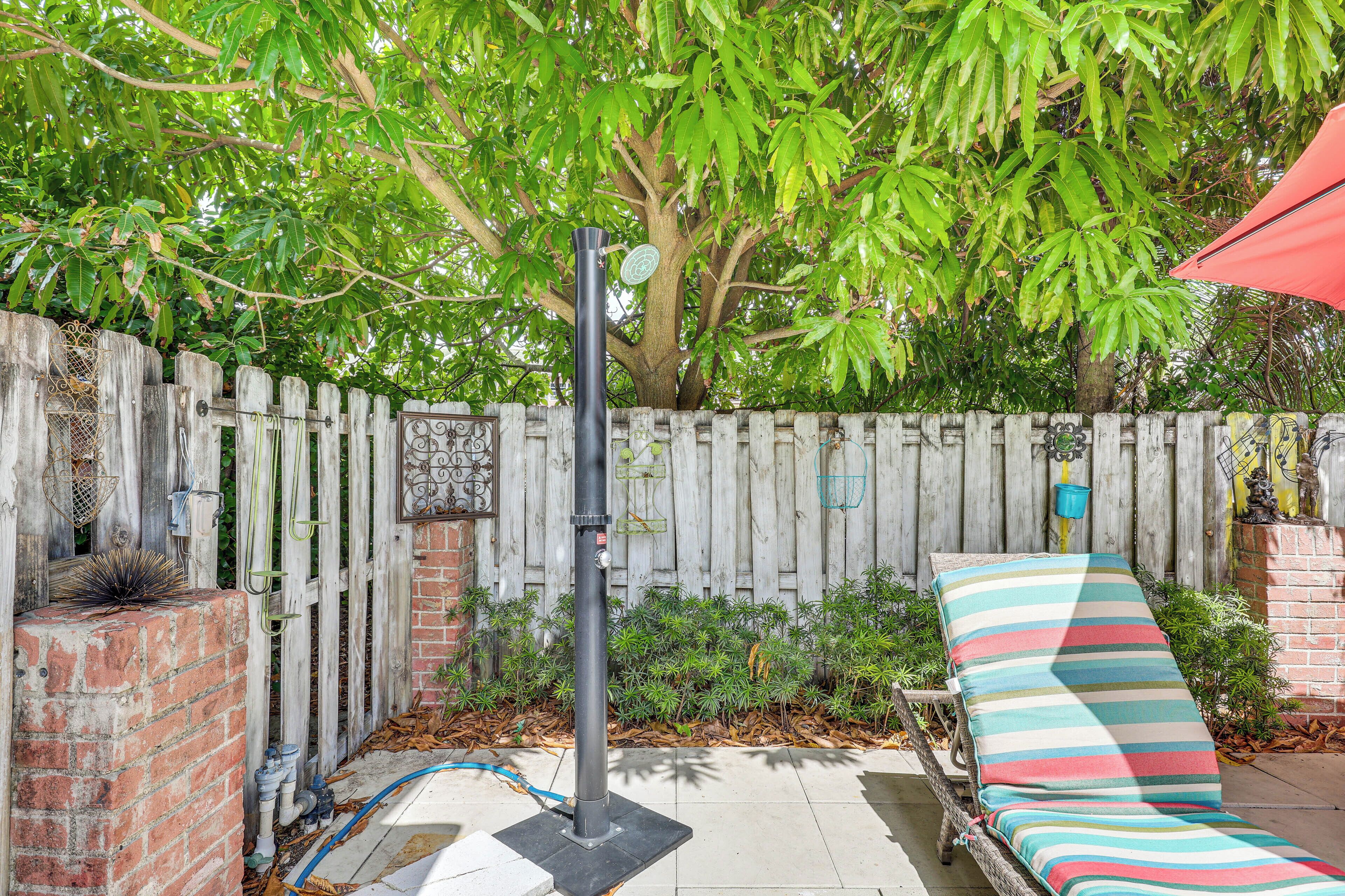 A backyard with a wooden fence, a brick wall, a metal pole, a lounge chair with a striped cushion, and a tree providing shade.