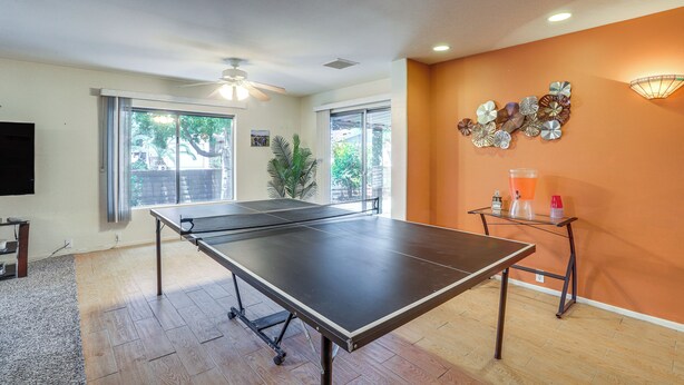A ping pong table in a room with large windows, a ceiling fan, and a wall-mounted light fixture.