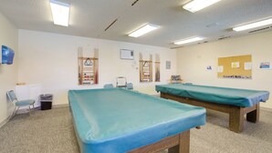 A room with two massage tables, a small chair, a trash bin, and a wall-mounted air conditioning unit.