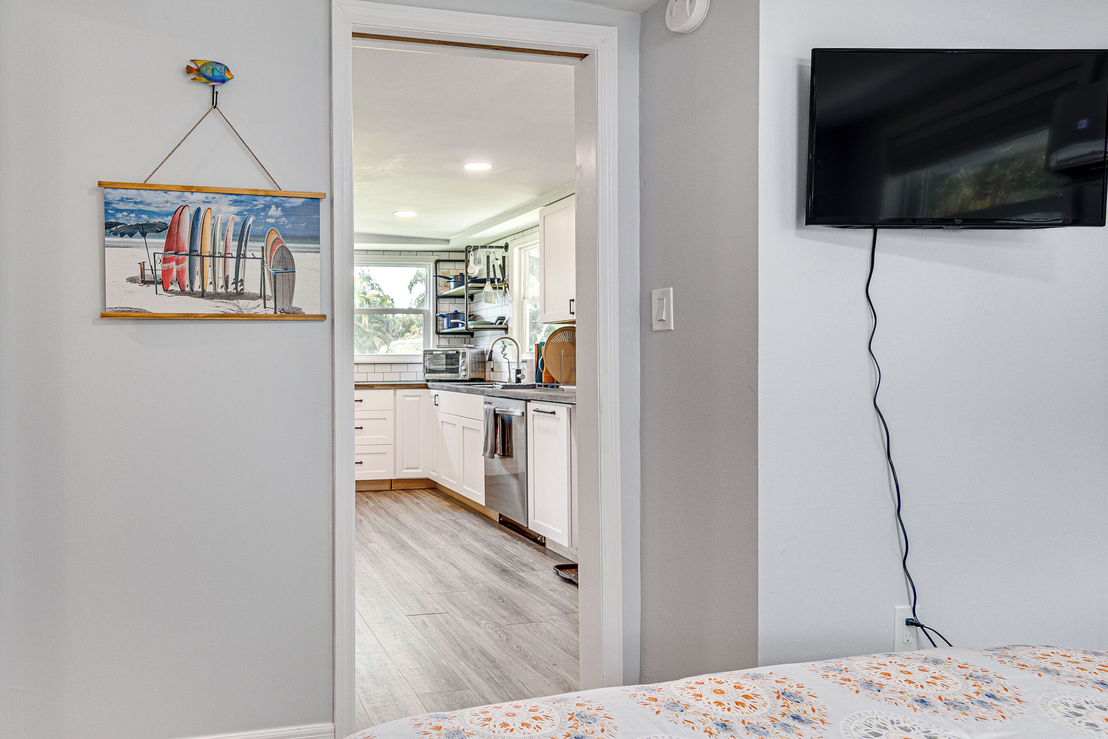 Una cocina moderna con armarios blancos, microondas y un lavamanos. Un cuadro de tablas de surf cuelga en la pared, y un televisor está montado allí.