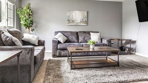 Una sala de estar con un sofá gris, una mesa de centro, una planta grande y un cuadro en la pared.