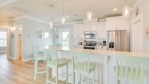 A modern kitchen with white cabinetry, a central island with bar stools, stainless steel appliances, and pendant lighting. - Hot Tub, Beach Access! Bay-view Gilchrist Retreat (Port Bolivar)
