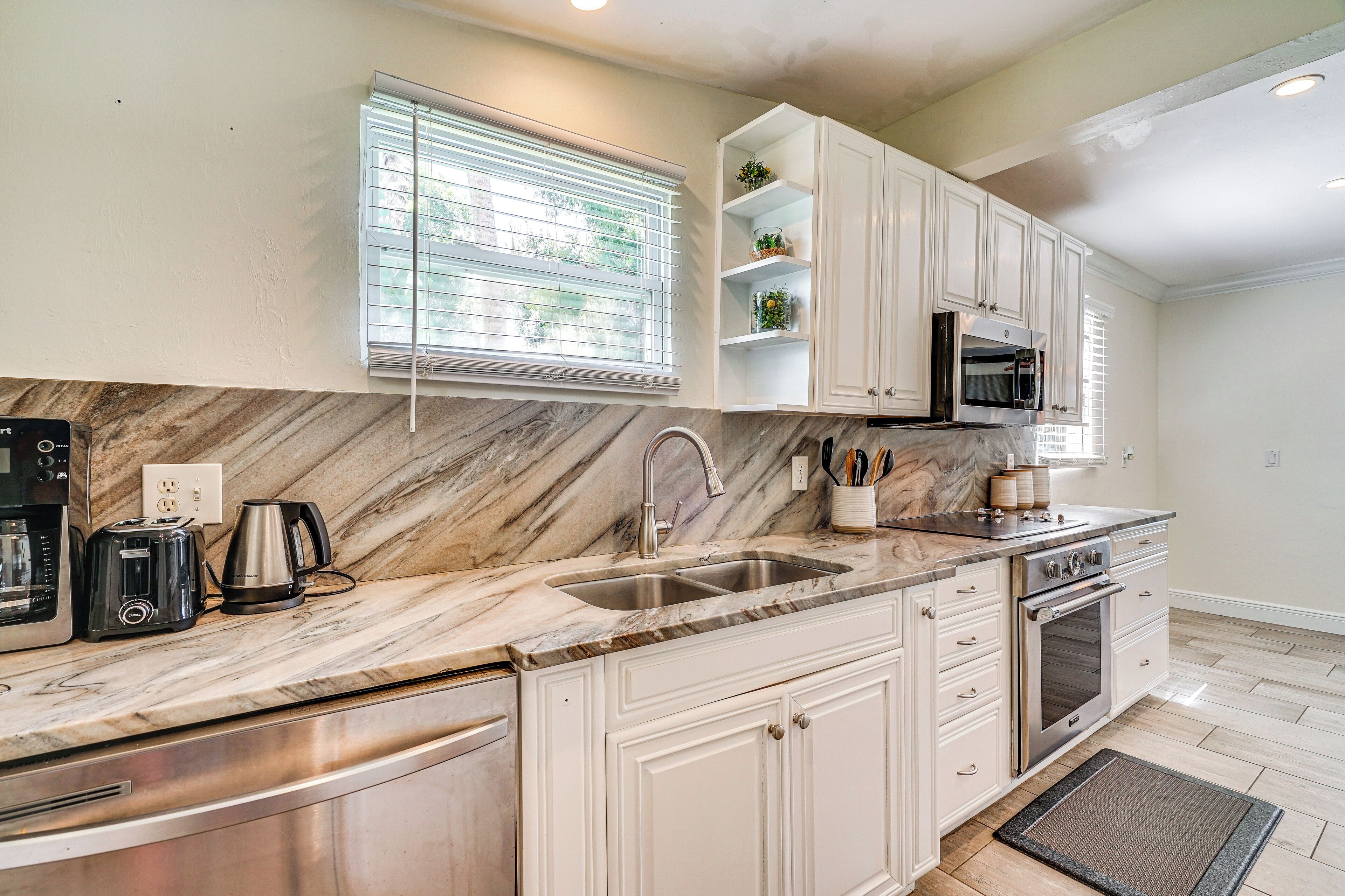 A modern kitchen with white cabinetry, a marble countertop, a built-in oven, and a window with blinds.