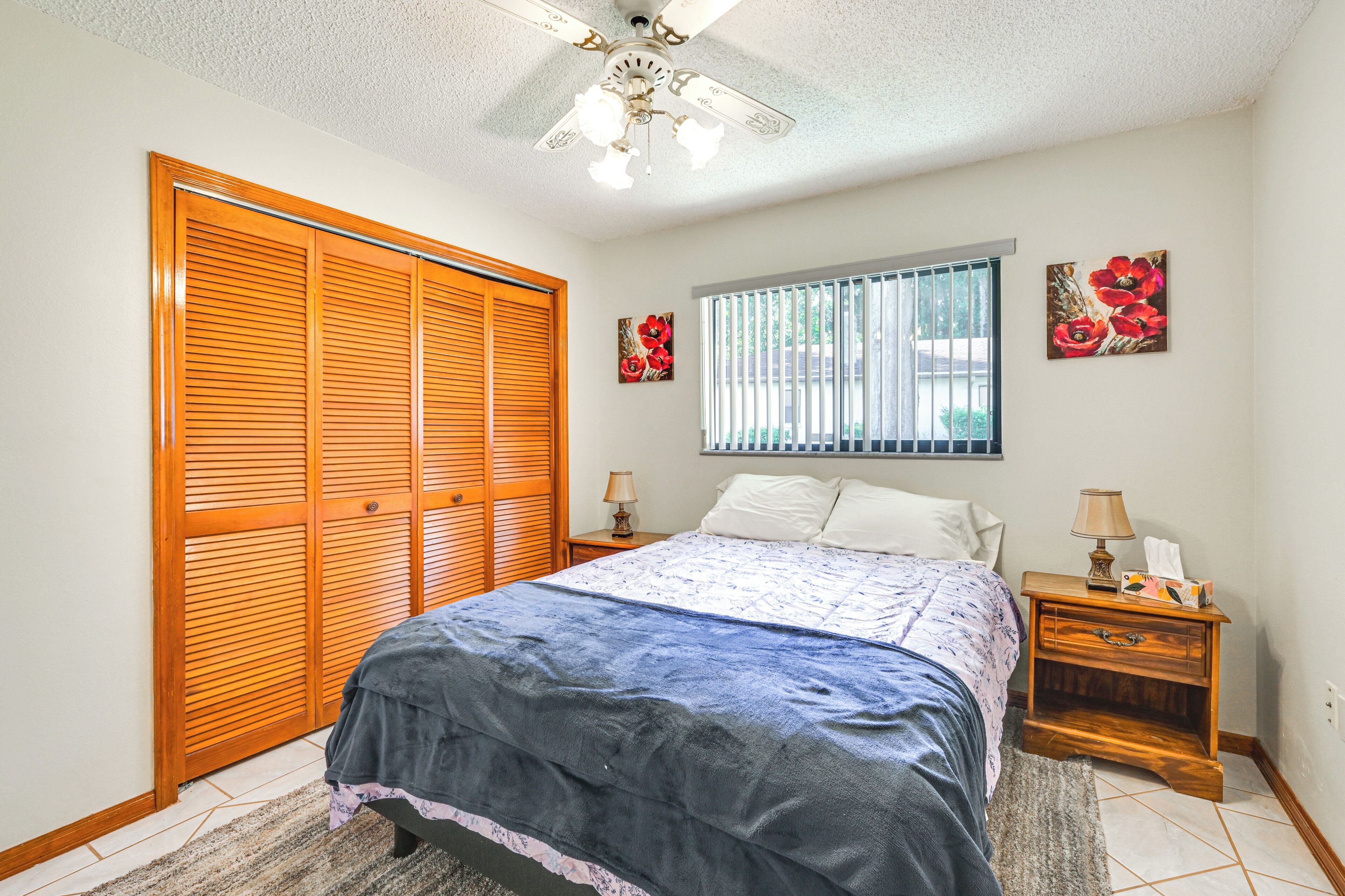A bedroom with a bed, wooden closet, window with blinds, nightstand, and two wall-mounted lamps.