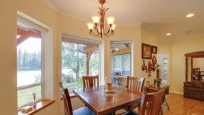 A dining area with a wooden table, chairs, a chandelier, and large windows offering a view of trees and a body of water. - Clark Fork Riverfront Home w/ Dock! (Thompson Falls)