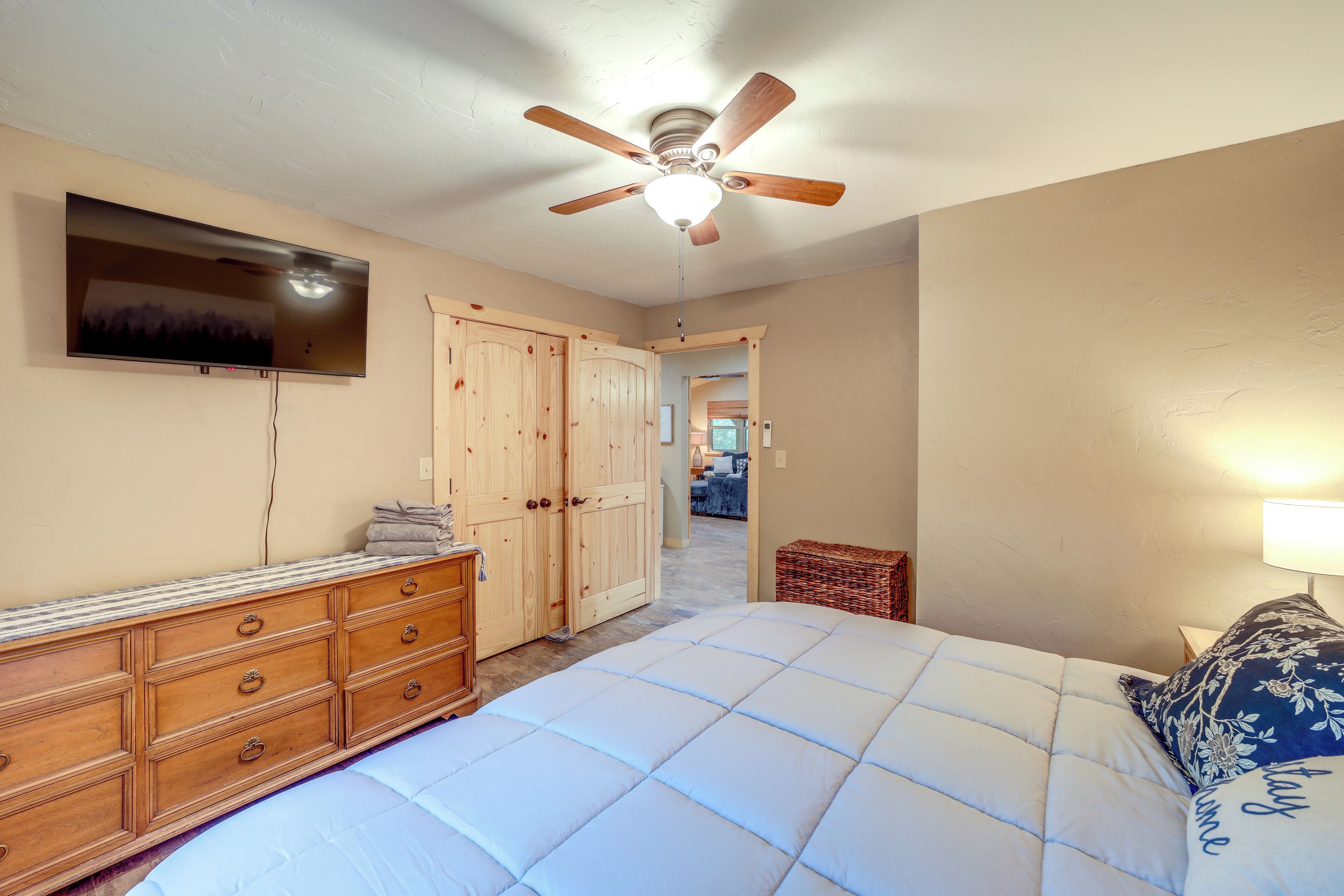 A bedroom with a bed, wooden dresser, TV, ceiling fan, and a hallway leading to another room.