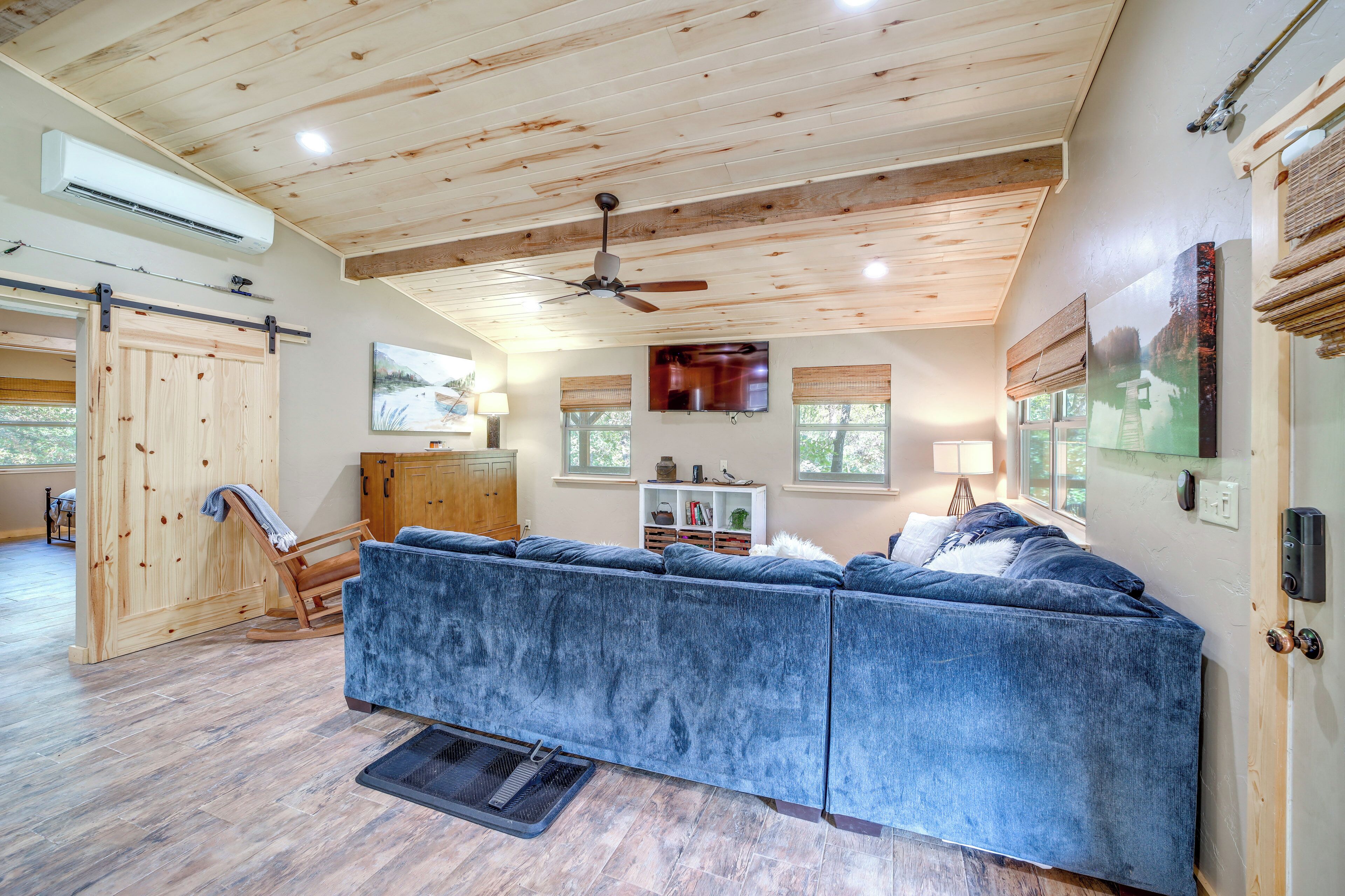Un vaste espace de vie agrémenté d’un grand canapé d’angle bleu, de poutres apparentes en bois au plafond, d’un ventilateur de plafond et d’une porte coulissante de style rustique.