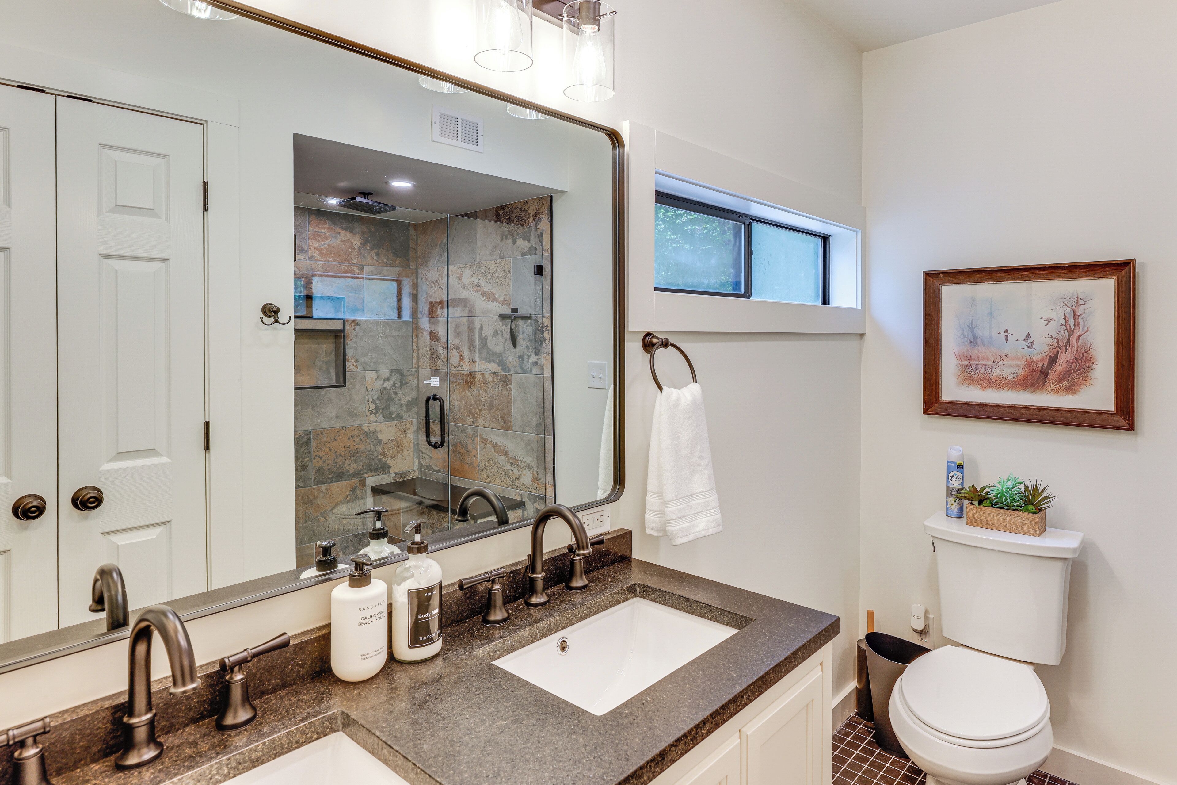 A bathroom with a large mirror, a sink with a granite countertop, a toilet, and a window above the sink.