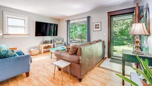 Un salon avec un canapé marron, un fauteuil bleu, une petite table blanche et une vue sur la verdure grâce à une grande fenêtre.