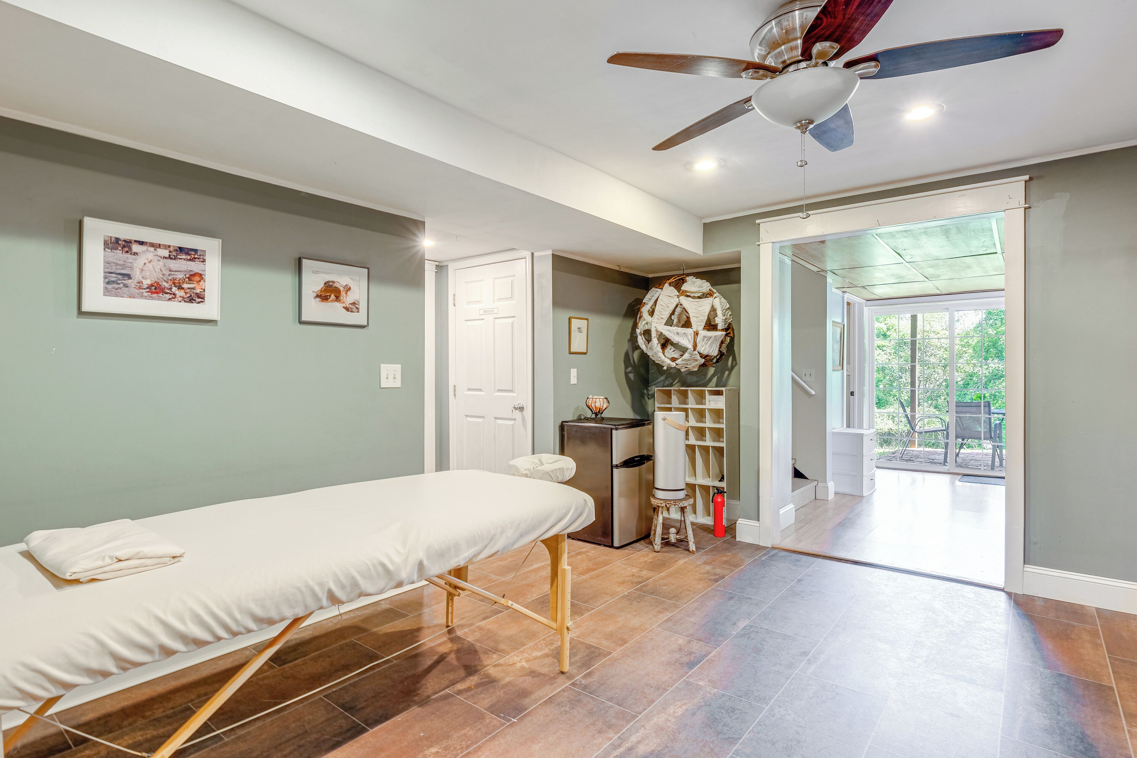 A room with a massage table, a ceiling fan, a door leading to a balcony, and a small refrigerator.