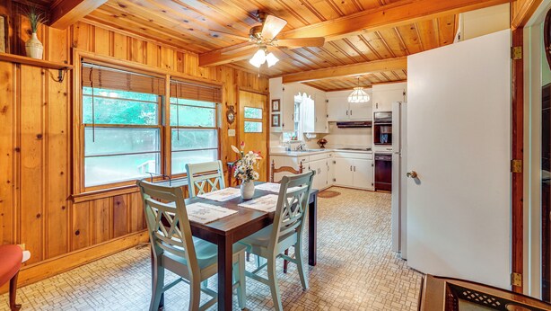 Interior de una cabaña de madera con una mesa de comedor, sillas, un ventilador de techo, una cocina con armarios blancos y un refrigerador.