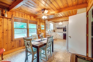 A wooden cabin interior with a dining table, chairs, a ceiling fan, a kitchen with white cabinets, and a refrigerator.
