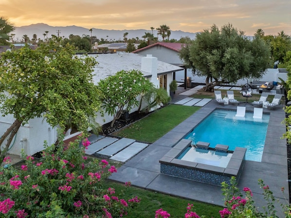 A swimming pool with a view of mountains, a covered patio, and a garden with pink flowers.
