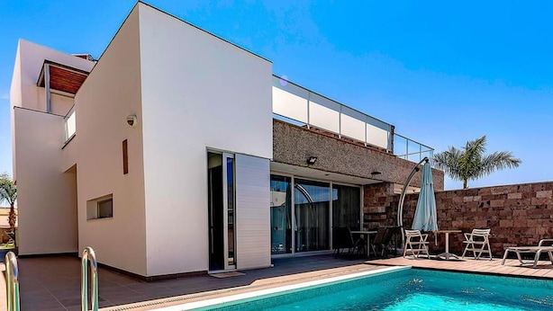 A modern two-story house with a swimming pool, outdoor seating, and a stone wall.