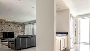 A modern living room with a stone wall, a flat-screen TV, a grey sofa, and a kitchen with white cabinets.