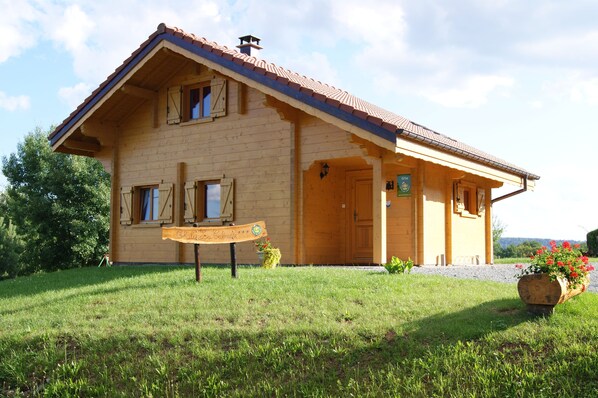 A wooden house with a tiled roof, a wooden sign in front, and a flower planter to the right.