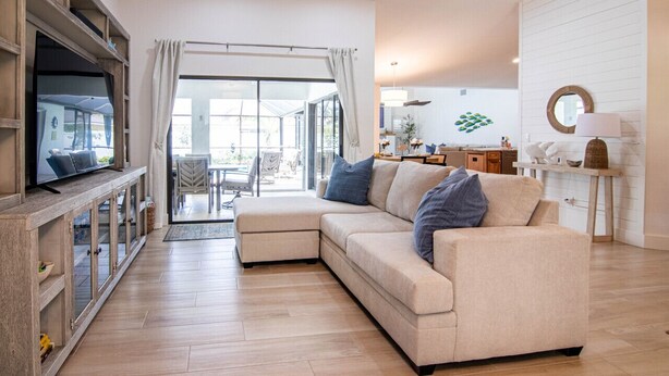 Una sala moderna con un sofá modular, un televisor sobre un mueble de madera, y un comedor visible a través de una puerta de cristal.