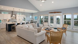 A spacious living area with a large sofa, wicker chairs, a dining table, and a kitchen with white cabinets and pendant lighting.