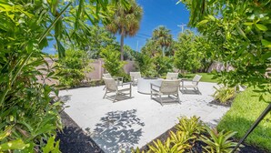 An outdoor patio with a round table and chairs, surrounded by greenery and a clear blue sky.