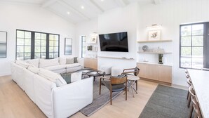 A spacious living room with a large sofa, a coffee table, and a TV mounted on the wall. There are windows providing natural light and a view of the outdoors. - Larkshine House at Lunara Bay (Key West)