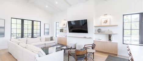 A spacious living room with a large sofa, a coffee table, and a TV mounted on the wall. There are windows providing natural light and a view of the outdoors.