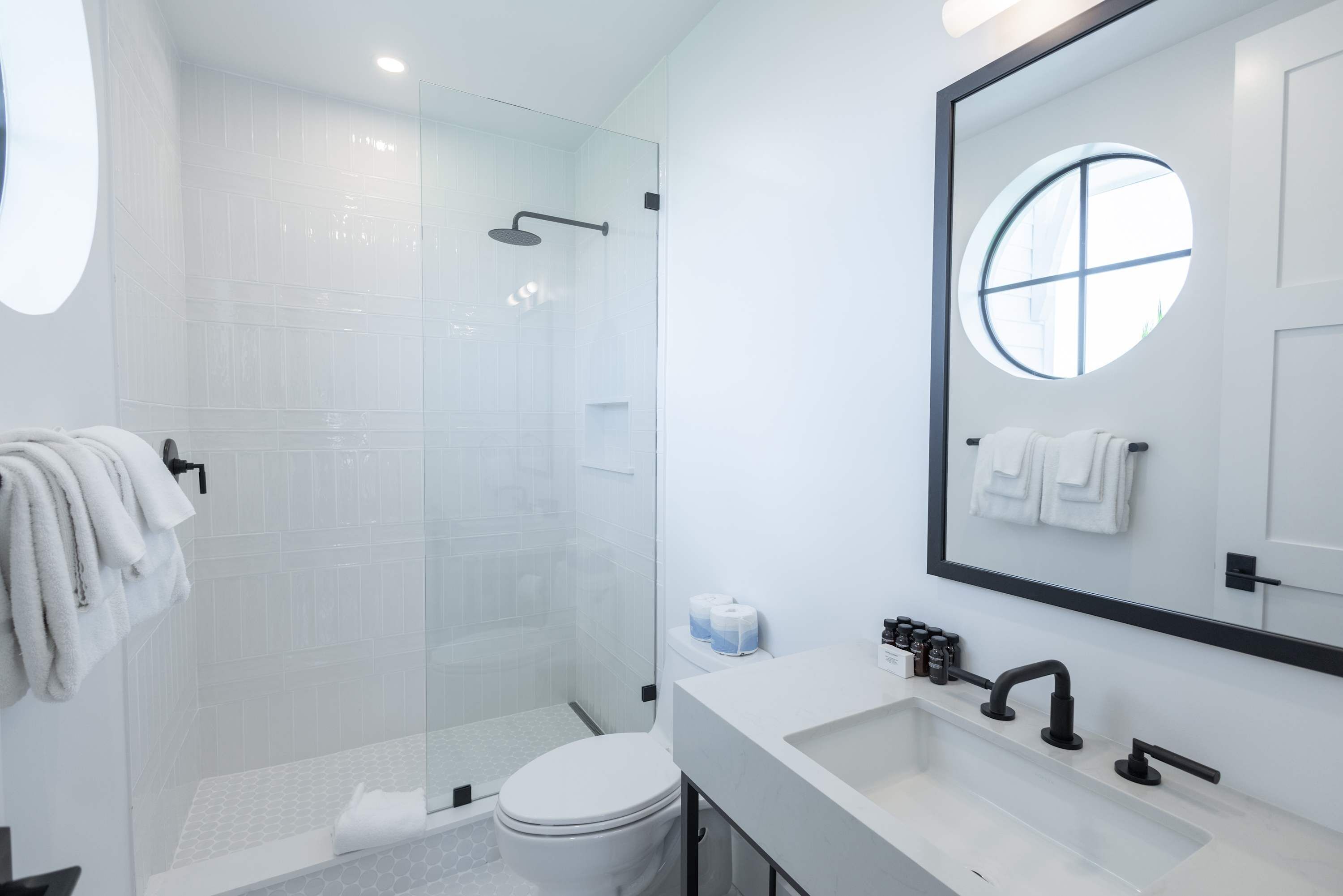 Une salle de bain moderne avec une douche à parois de verre, un lavabo blanc et un grand miroir orné d’une fenêtre ronde.