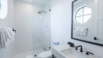 Une salle de bain moderne avec une douche à parois de verre, un lavabo blanc et un grand miroir orné d’une fenêtre ronde.