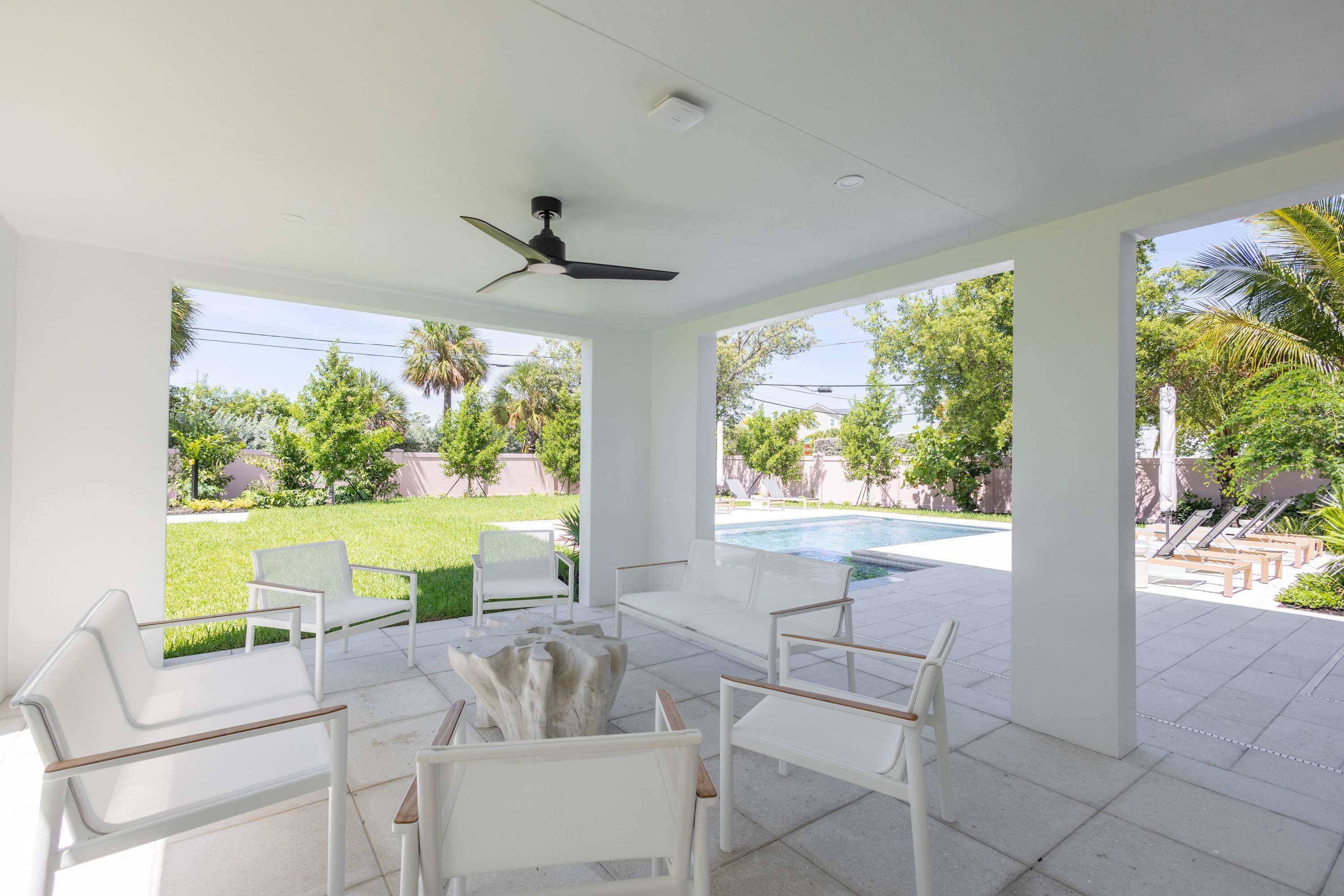 A covered outdoor seating area with white furniture, a ceiling fan, and a view of a swimming pool and greenery.
