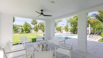 A covered outdoor seating area with white furniture, a ceiling fan, and a view of a swimming pool and greenery.