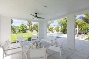 A covered outdoor seating area with white furniture, a ceiling fan, and a view of a swimming pool and greenery. - Larkshine House at Lunara Bay (Key West)