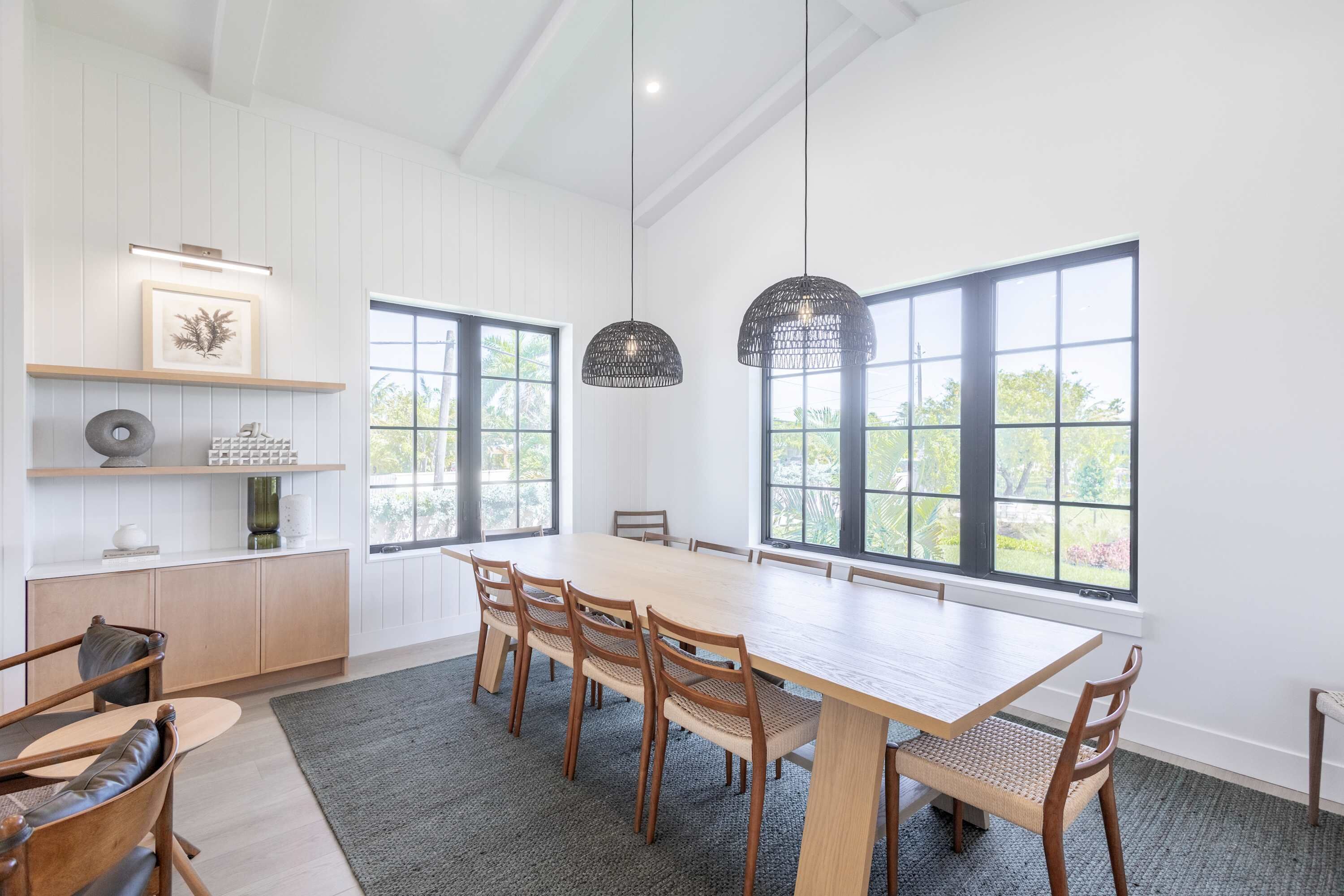 A spacious dining area with a long table, multiple chairs, and large windows offering a view of greenery.