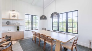 A spacious dining area with a long table, multiple chairs, and large windows offering a view of greenery.