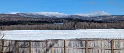 A snowy landscape with a wooden fence in the foreground and a mountain range in the background.