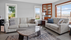 A modern living room with two sofas, a coffee table, and a side table. There are large windows providing a view of the outdoors. - Stowe Retreat Mountain Views & Fenced Backyard by RedAwning (Stowe)