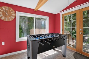 A room with a foosball table, a wooden clock, and a window with a view of greenery. - Stowe Retreat Mountain Views & Fenced Backyard by RedAwning (Stowe)