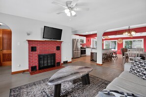 A modern living room with a fireplace, a television mounted above it, and a dining area with a table and chairs. - Stowe Retreat Mountain Views & Fenced Backyard by RedAwning (Stowe)