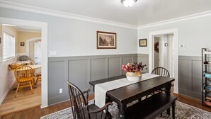 A dining area with a dark wooden table, white walls, and a gray wainscoting. There are wicker chairs and a bench around the table, which has a vase of flowers. A bookshelf is visible to the right. - Charming Downtown Mccloud Retreat 2 Bedroom Home by RedAwning (McCloud)