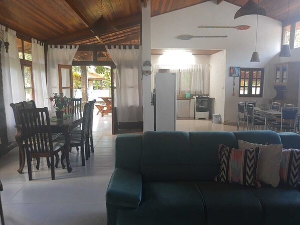 A spacious living area with a green sofa, wooden dining table, and chairs. There is a kitchen area with a refrigerator and a dining table.