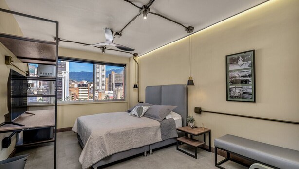 A modern bedroom with a bed, desk, and bench, featuring a city view through a large window.
