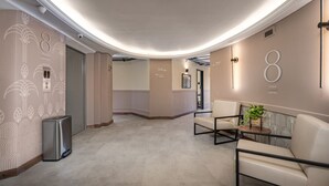 Un pasillo moderno con techo curvo, un cubo de basura, una zona de estar con mesa y planta, y señales direccionales que indican los niveles.