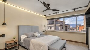 Un dormitorio moderno con una cama grande, un banco, una mesita de noche, un ventilador de techo y una ventana con vistas a la ciudad.
