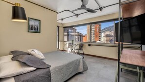 A bedroom with a bed, a desk, and a chair, a ceiling fan, and a large window with a city view.