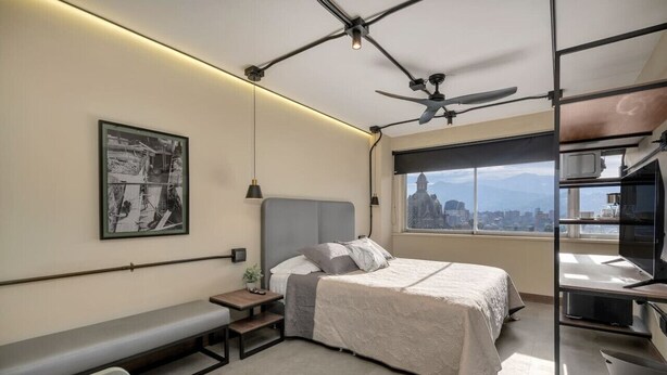 Un dormitorio moderno con cama, banco, obras de arte y vista a la ciudad.