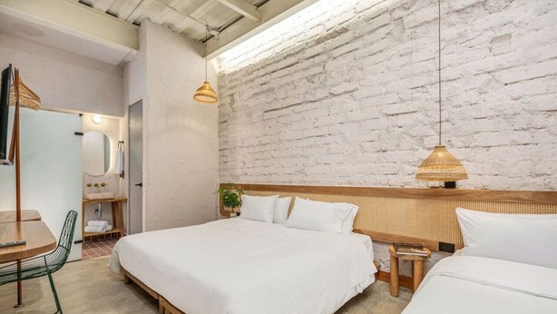 Une chambre moderne avec un mur en briques blanches, deux lits, une tête de lit en bois, une lampe suspendue et un petit tabouret.