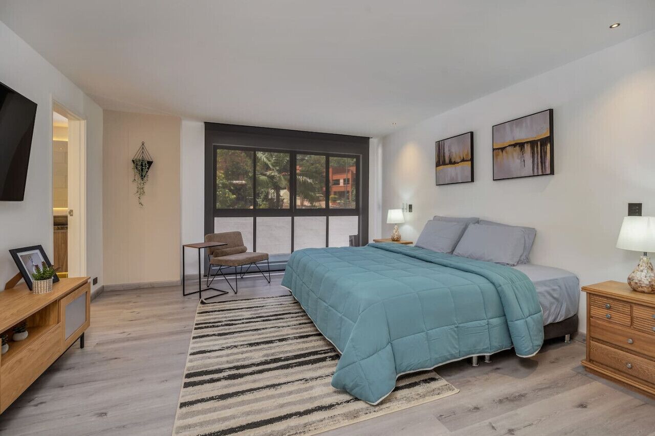 A modern bedroom with a large bed, a desk, and a chair. There is a window with a view of buildings outside.