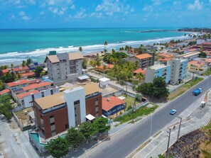 An aerial view of a coastal residential area with buildings, a road, and a beach. - Flat Zero7 Villa del Porto com piscina  (Ipojuca)