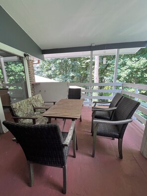 Ein überdachter Patio-Bereich mit Korbmöbeln, einem Holztisch und Blick auf Bäume.